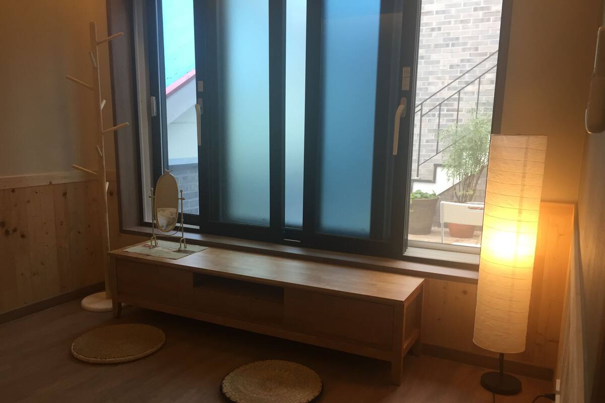 Une pièce avec une grande fenêtre, un banc en bois, une petite table avec un miroir et une lampe.