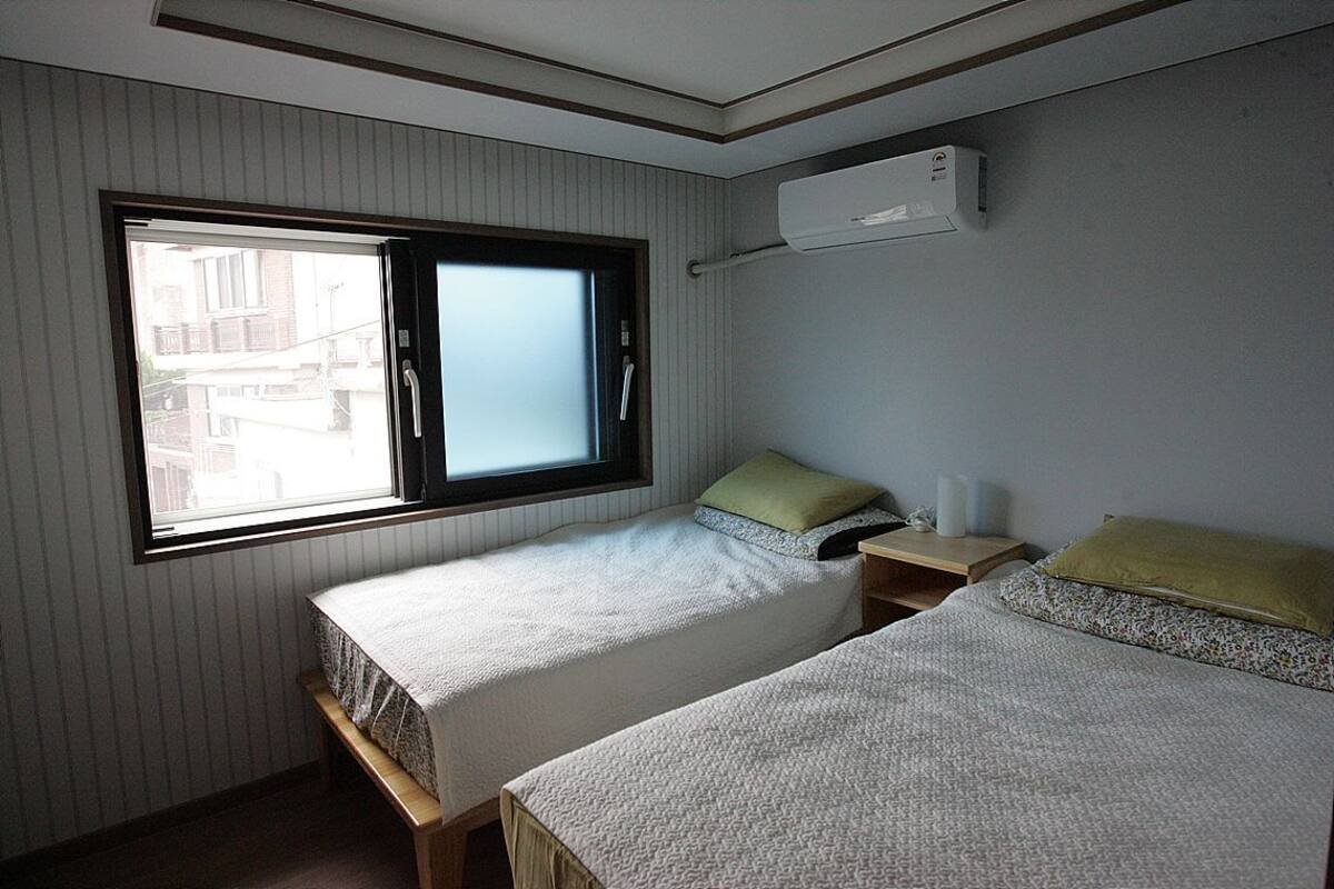 Une chambre avec deux lits, une fenêtre, un climatiseur et une table de chevet avec une lampe.