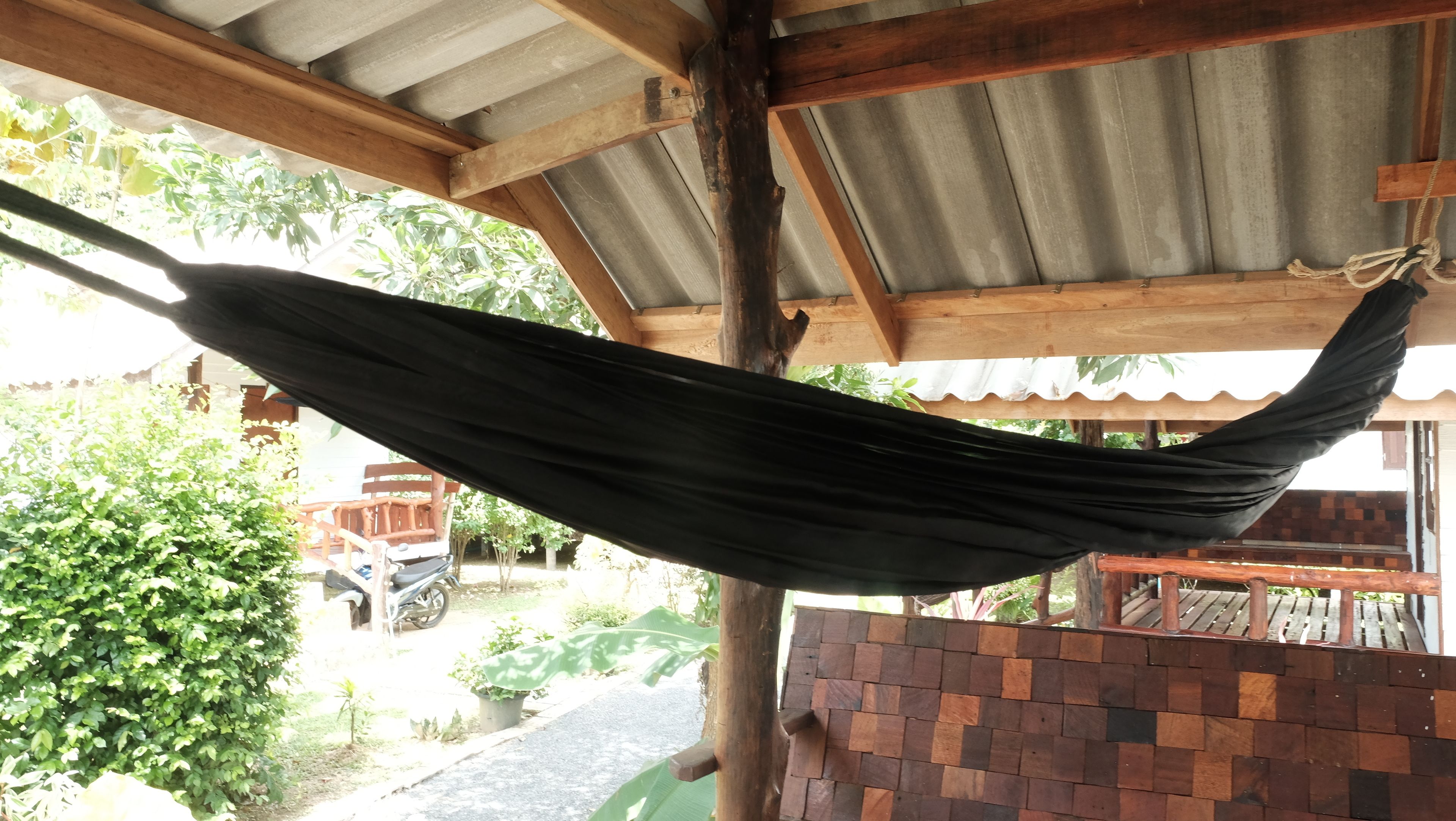 A hammock hanging under a wooden structure with a corrugated metal roof, surrounded by greenery and a brick wall.