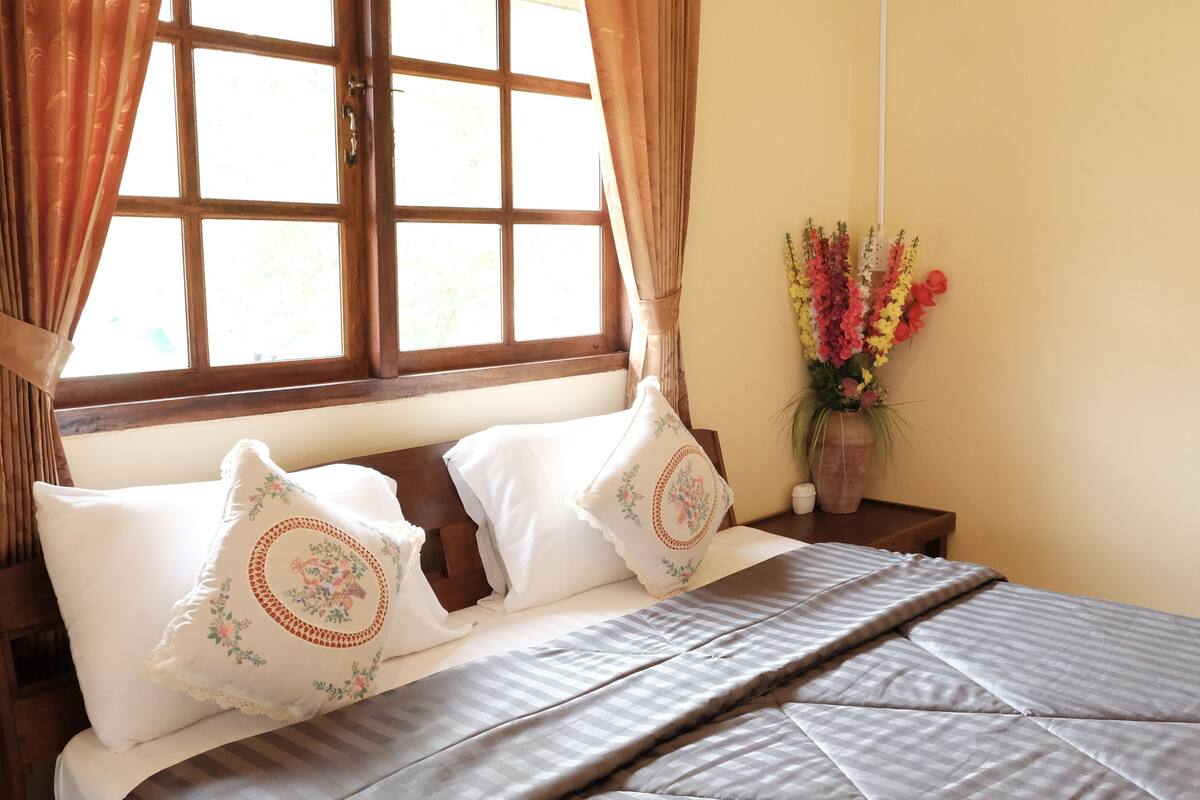 Una cama bien hecha con dos almohadones decorativos, un cabecero de madera y una ventana con cortinas.