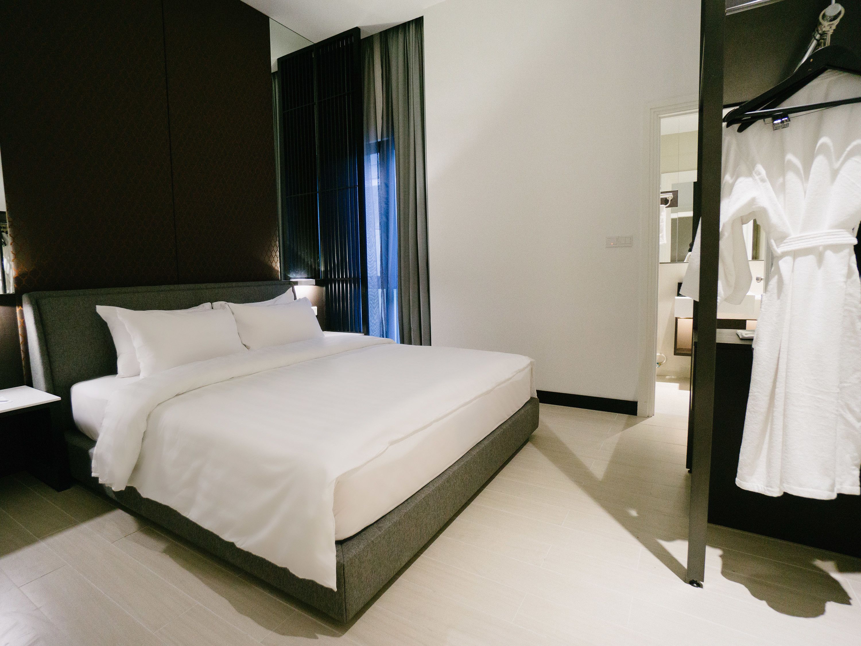 A modern hotel room with a large bed, a bedside table, a wardrobe, and a bathroom visible through an open door.