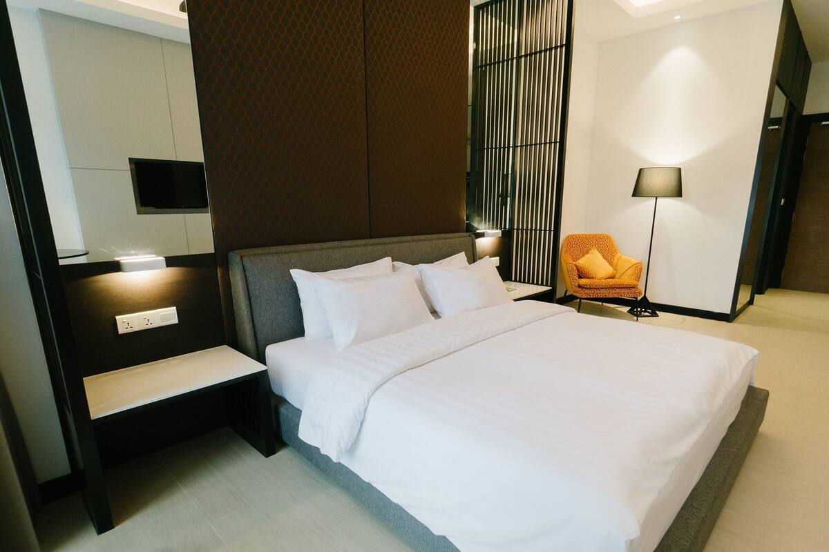 A modern hotel room with a large bed, a bedside table, a chair, and a lamp.