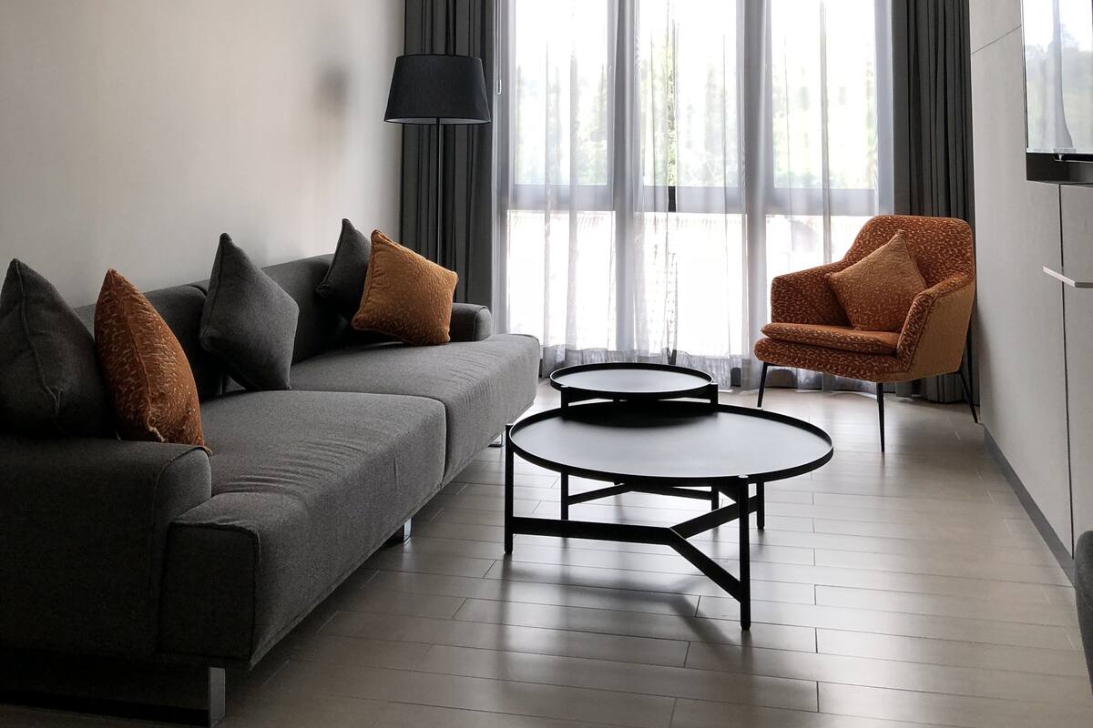 A modern living room with a grey sofa, a coffee table, and an orange armchair. There is a ceiling fan, a TV mounted on the wall, and a window with curtains.