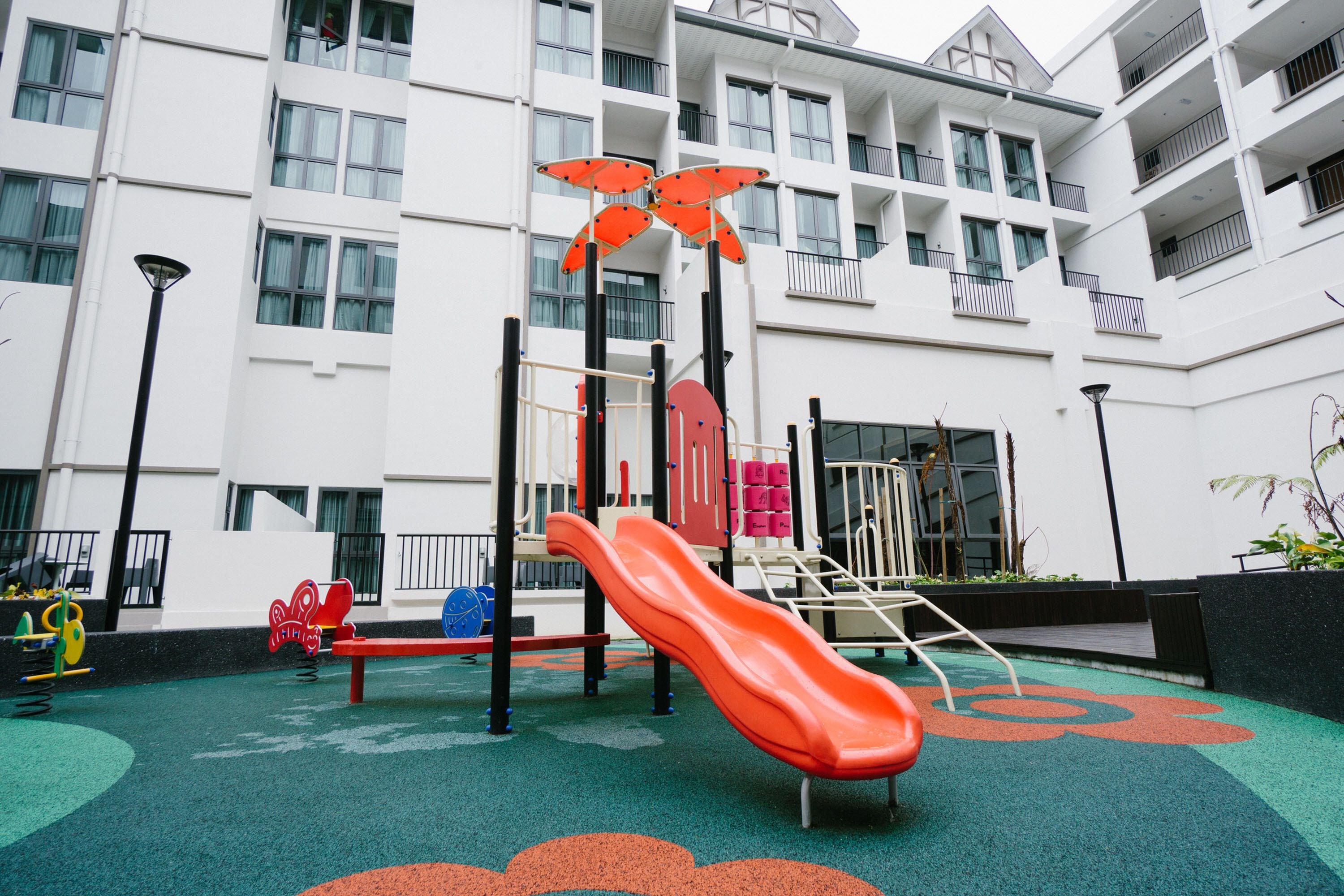 A playground with a red slide and various play equipment, surrounded by a white building with balconies.