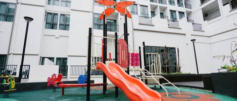A playground with a red slide and various play equipment, surrounded by a white building with balconies.