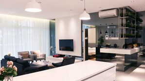 A modern living room with a white sofa, a coffee table, and a TV. There is a kitchen area with a white countertop and a dining table.