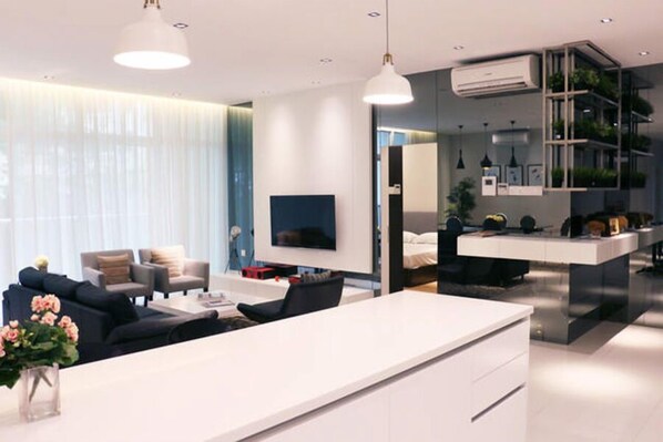 A modern living room with a white sofa, a coffee table, and a TV. There is a kitchen area with a white countertop and a dining table. - Sembilan (George Town)