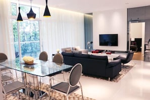 A modern living room with a glass dining table, dark sofas, a flat-screen TV, and large windows with curtains.