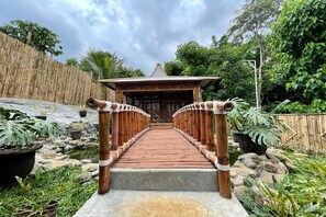 A wooden bridge leading to a traditional wooden structure with a tiled roof, surrounded by lush greenery and a bamboo fence. - Rizky Bromo Homestay (Sukapura)