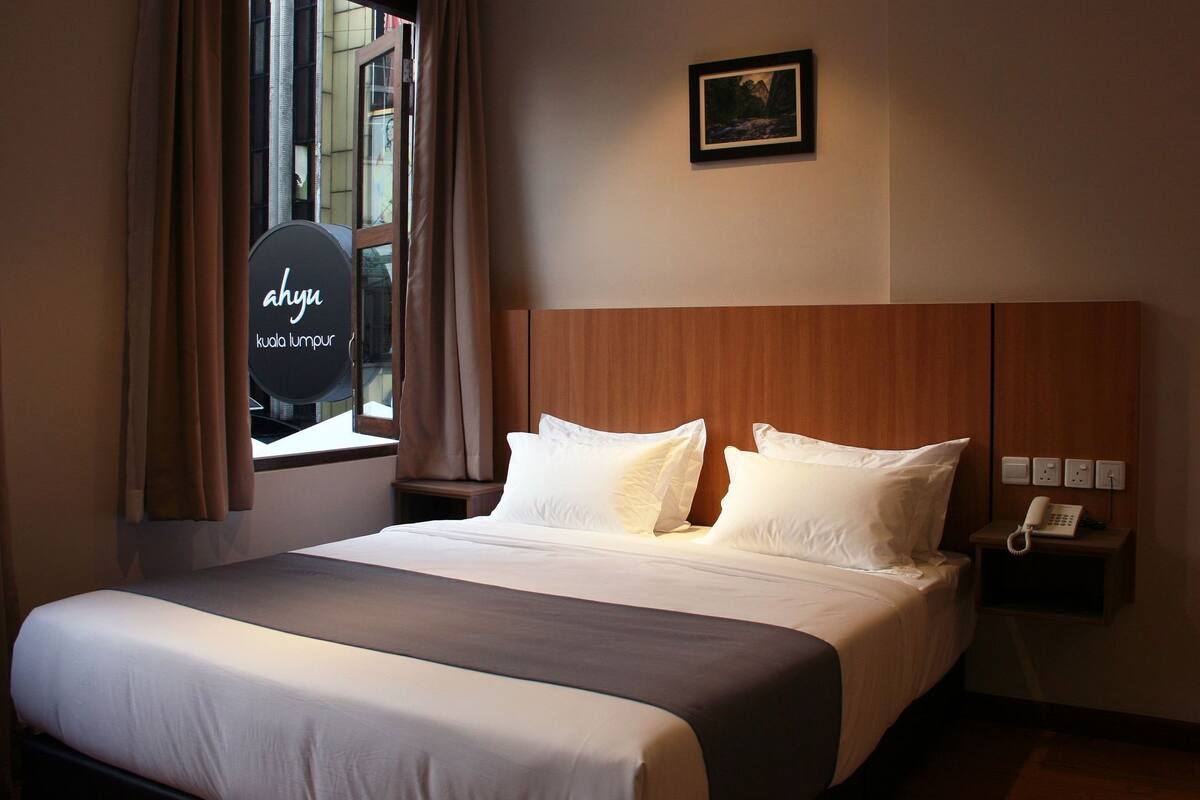 A hotel room with a bed, a nightstand with a phone, and a window with curtains.