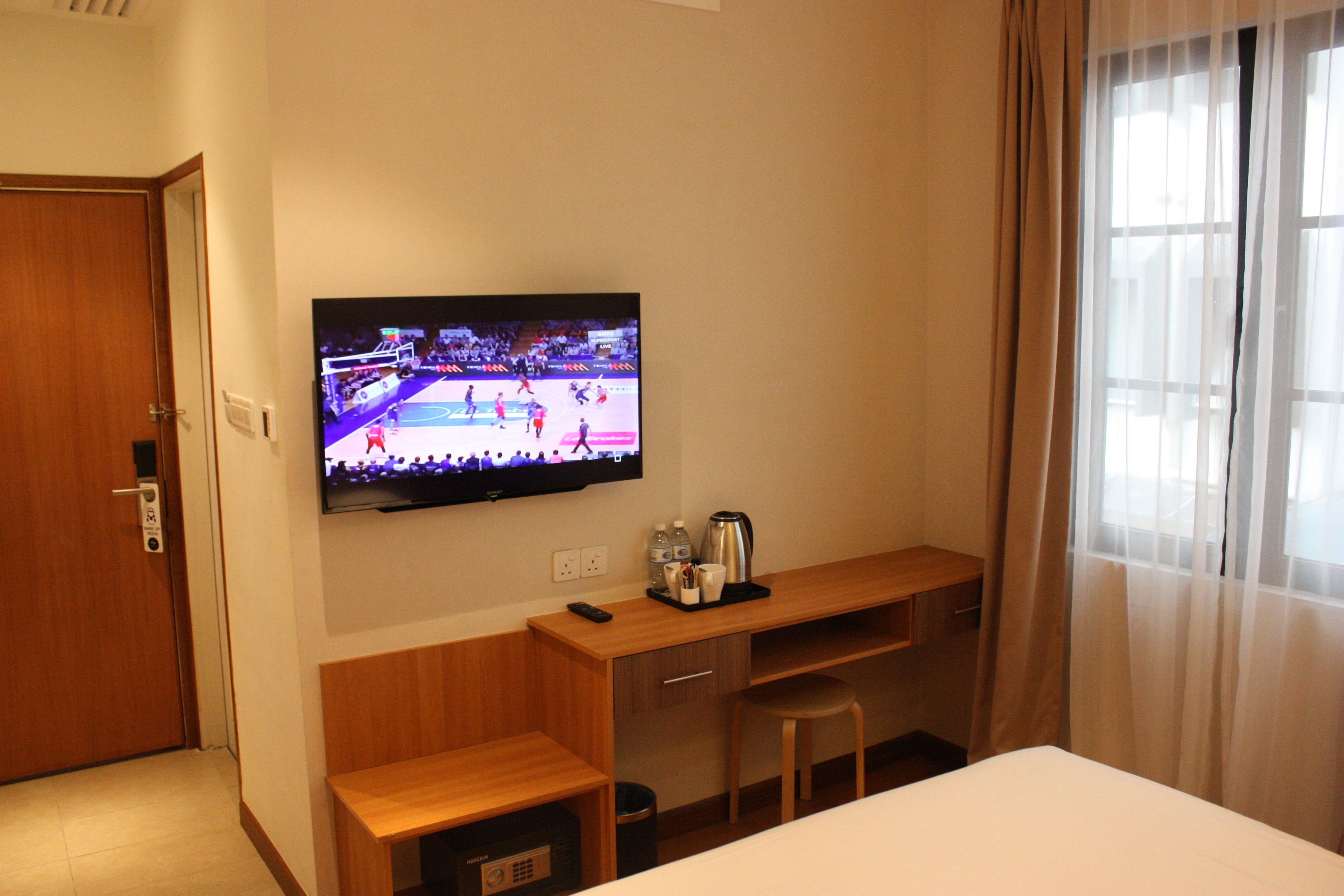 Habitación de hotel con televisor de pantalla plana montado en la pared, un escritorio de madera con control remoto, una cafetera y un cesto de basura.