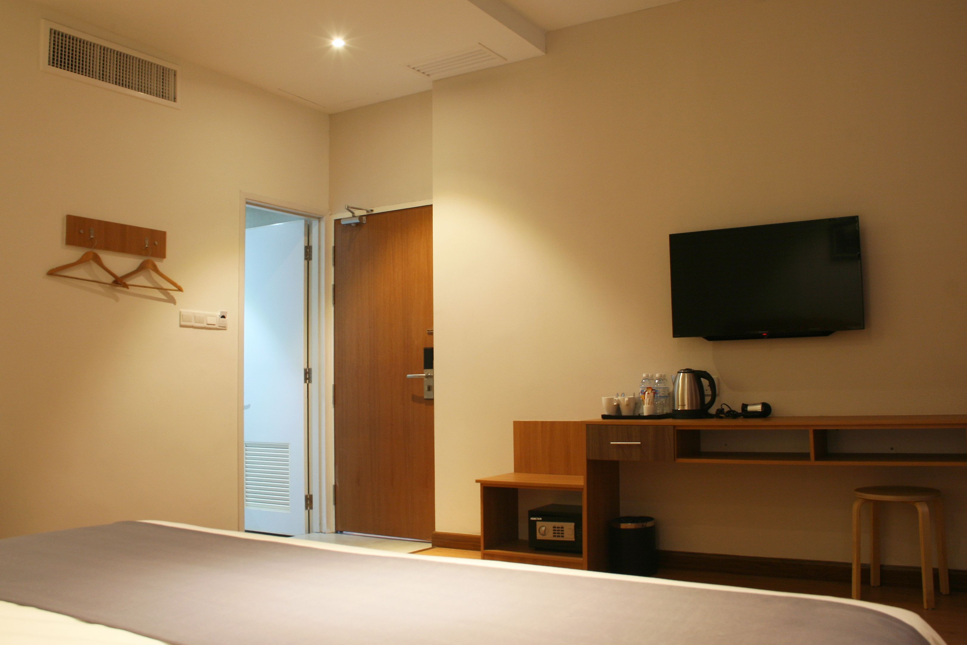 A hotel room with a bed, a television mounted on the wall, a wooden desk with a kettle, and a small stool.