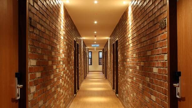 Un pasillo largo y estrecho con paredes de ladrillo y puertas de madera, que conduce a una salida luminosa.
