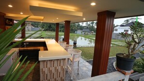A modern outdoor bar area with a wooden counter and stools, overlooking a rice paddy field. - Darra Ria Villa Ubud (Ubud)