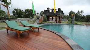 A wooden deck with two lounge chairs, a swimming pool, and a temple-like structure with flags. - Darra Ria Villa Ubud (Ubud)