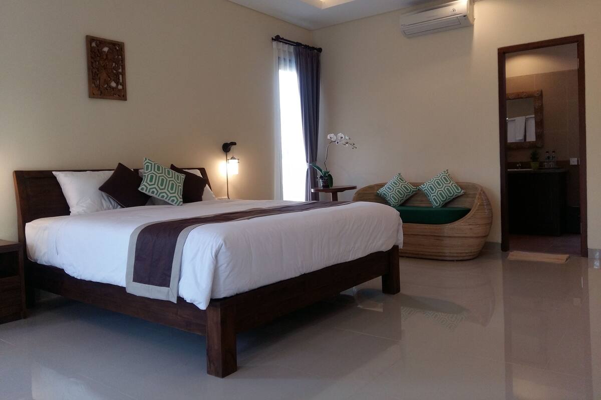 A well-lit bedroom with a large bed, a bedside table, a lamp, and a window with curtains.