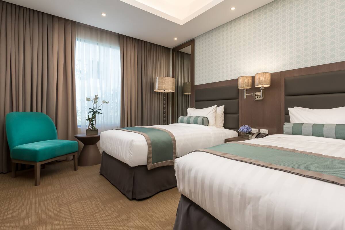 Ein Hotelzimmer mit zwei Betten, einem türkisfarbenen Sessel, einem kleinen Tisch mit Pflanze und einem großen Fenster mit Vorhängen.