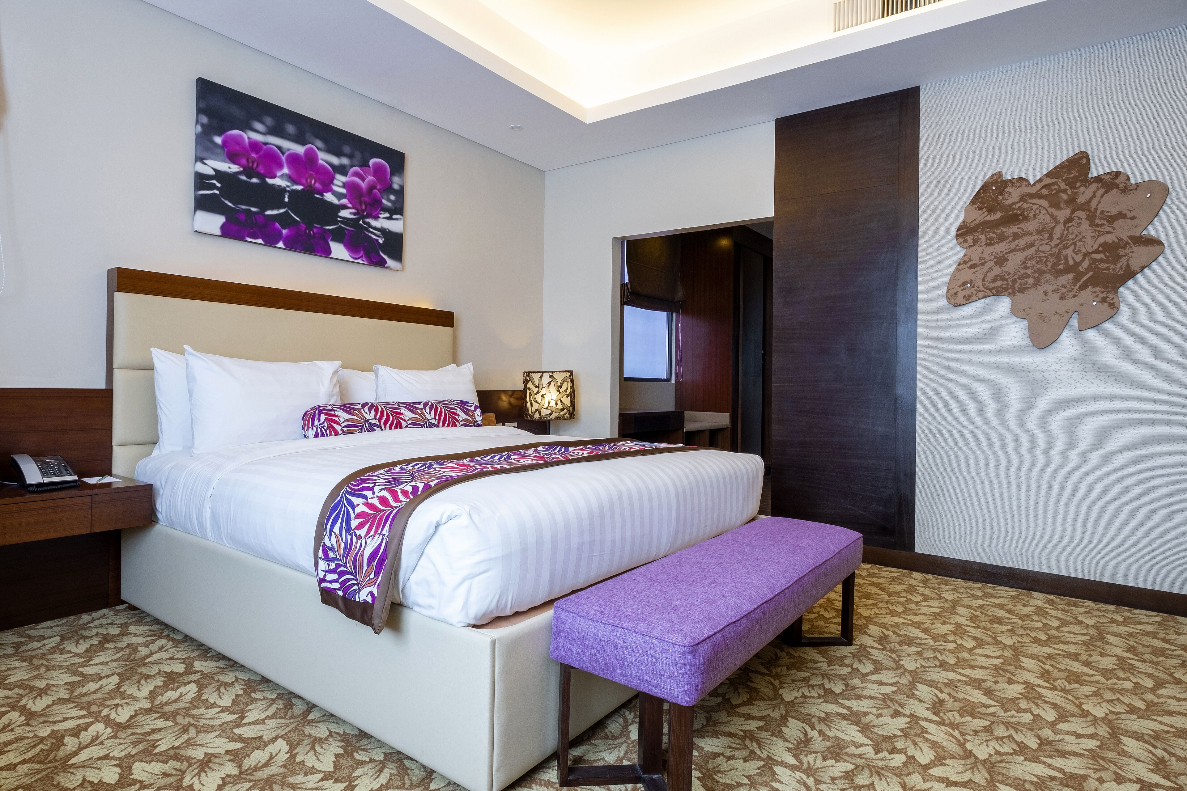 A hotel room with a large bed, a bedside table with a lamp, a bench, and a wall with a floral painting.