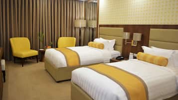 A hotel room with two beds, a yellow armchair, a small table with a vase of flowers, and a lamp.