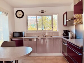 A modern kitchen with stainless steel appliances, a window with a view of trees, and a wall clock. - Villa with sea view, heated swimming pool  (Groix)
