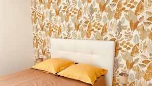 Una cama con cabecera blanca y dos almohadas amarillas, una mesita de noche con lámpara, y una pared con diseño de hojas.