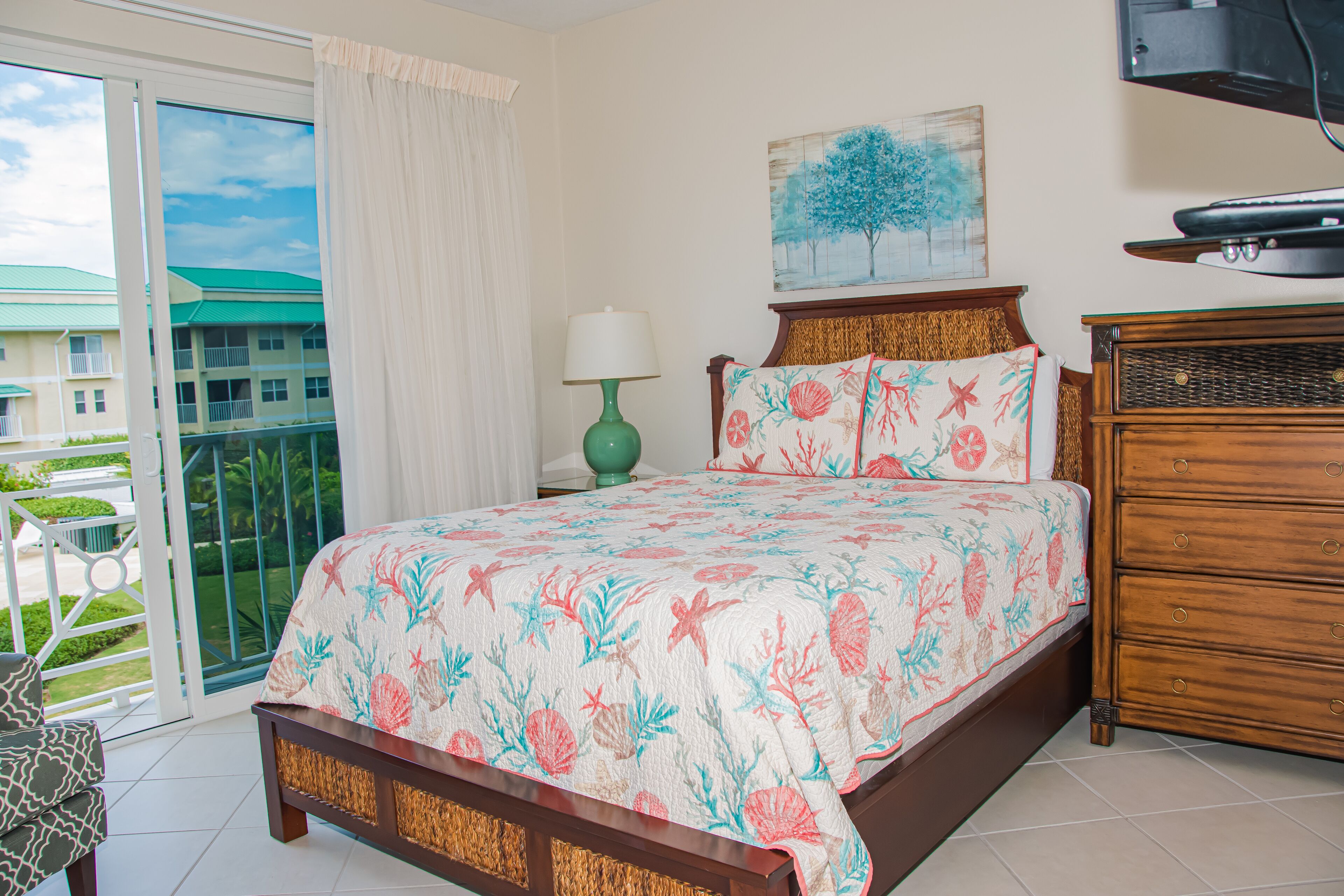 A bedroom with a bed, a dresser, a lamp, a painting, and a view of the outside.