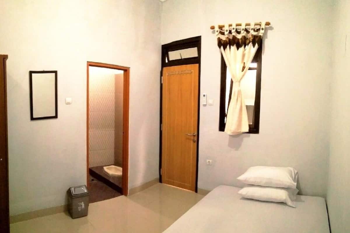 Un dormitorio con cama, puerta, un cubo de basura pequeño y ventana con cortinas.