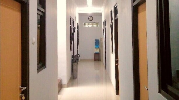 A long, narrow hallway with doors on both sides, a clock on the wall, and a trash can in the center. - Adikarta Guesthouse Janti (Banguntapan)