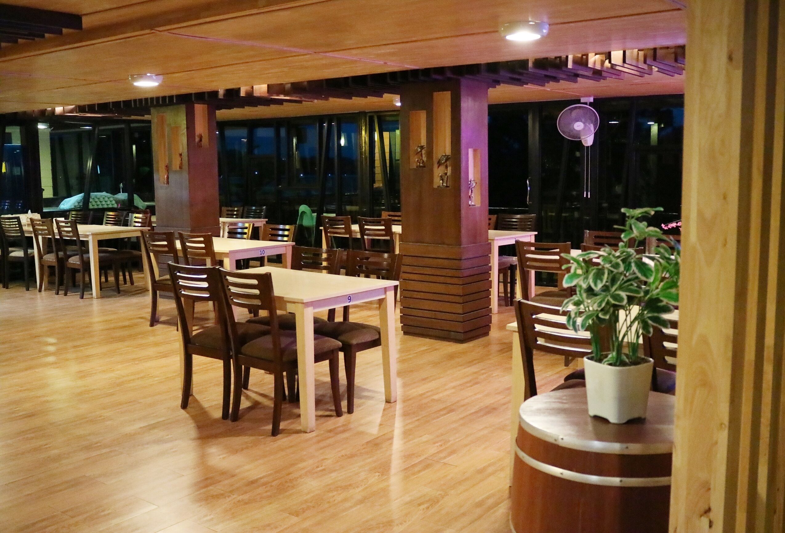 A restaurant with wooden tables and chairs, large windows, and a fan.
