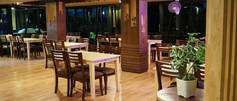 A restaurant with wooden tables and chairs, large windows, and a fan.