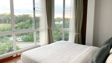 Un dormitorio con un ventanal grande, una cama con ropa de cama blanca y vistas a zonas verdes y edificios.