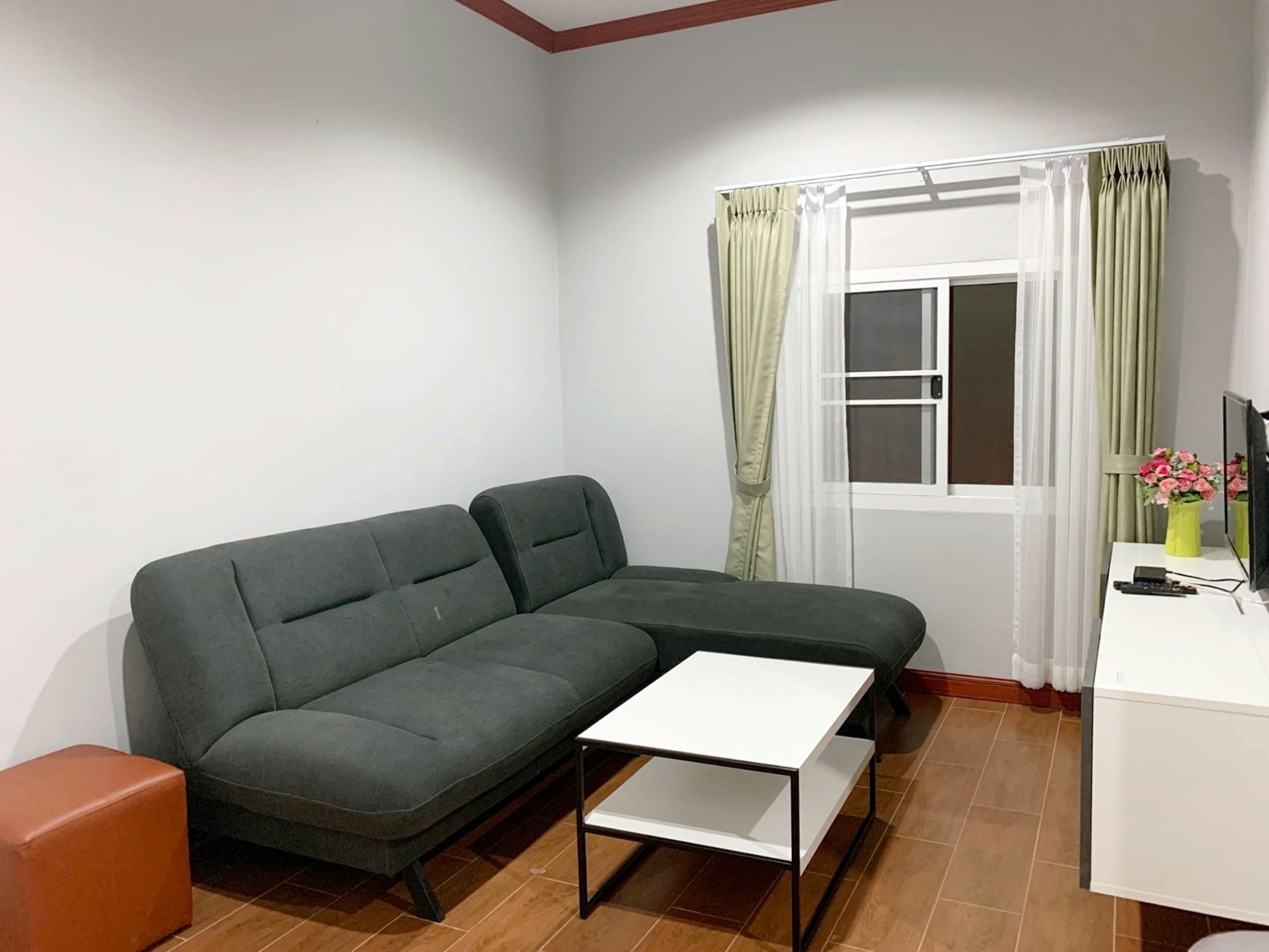 A living room with a sectional sofa, a small white table, a TV, and a window with curtains.