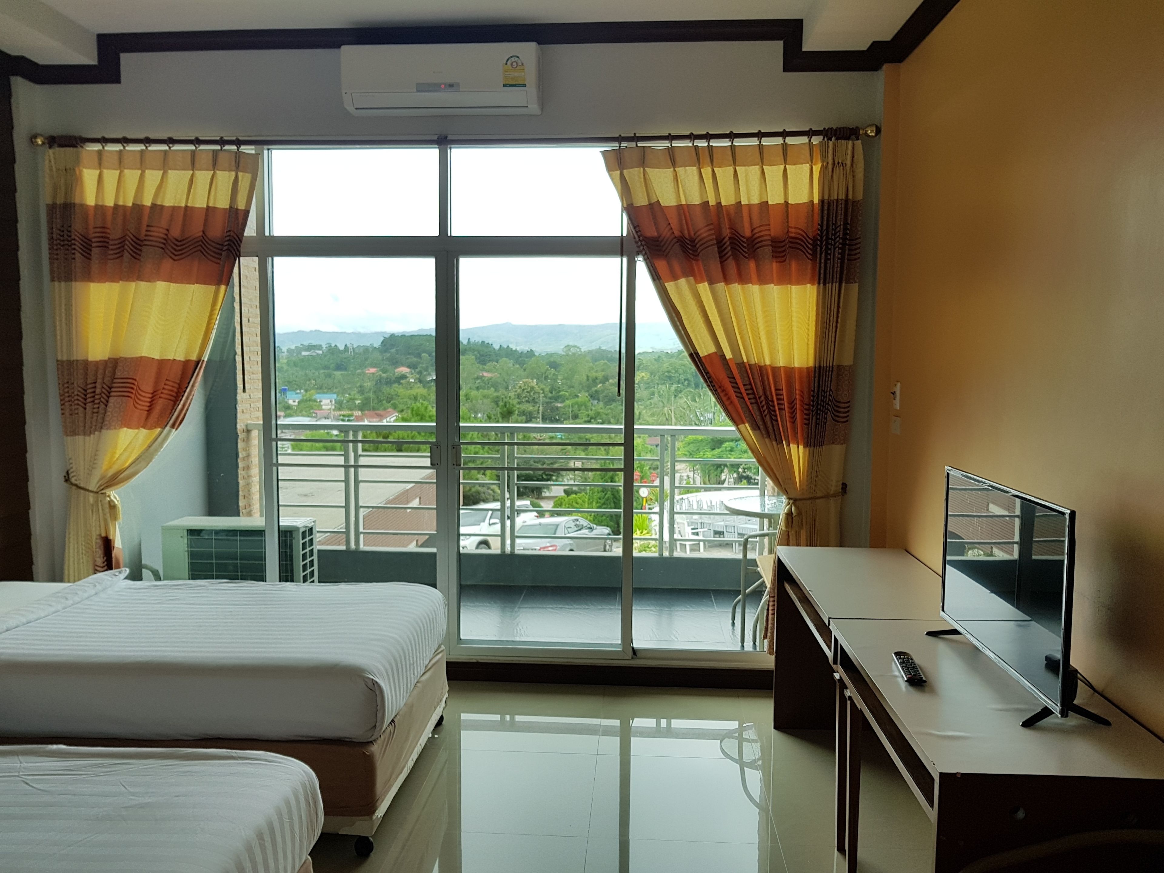 A hotel room with two beds, a TV, and a balcony with a view of the city and mountains.