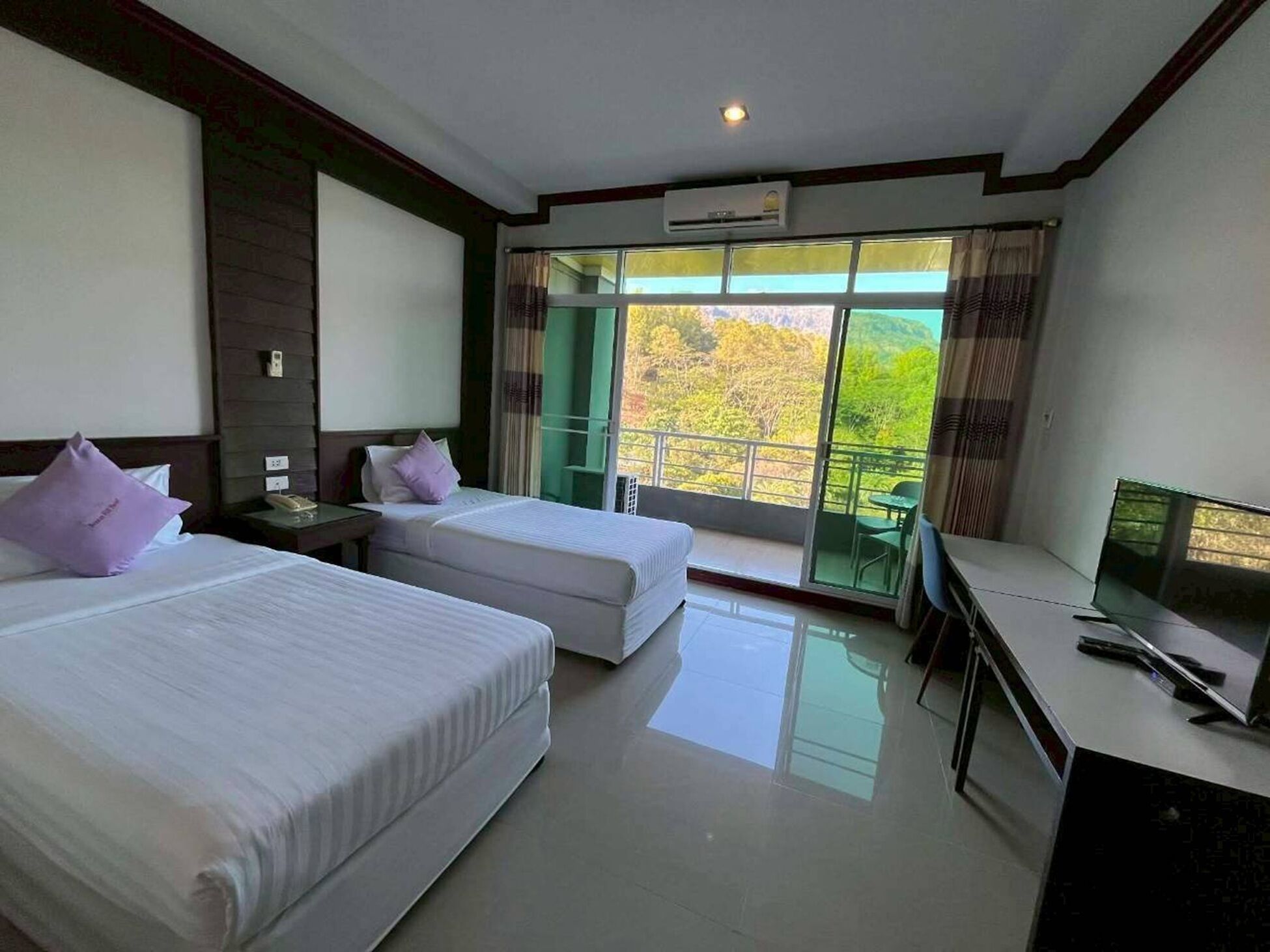 Habitación de hotel con dos camas, un escritorio con televisor y un balcón con vistas a árboles.