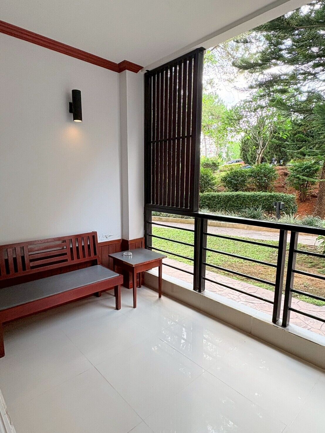A balcony with a wooden bench and a small table, a black metal railing, and a view of a green garden.