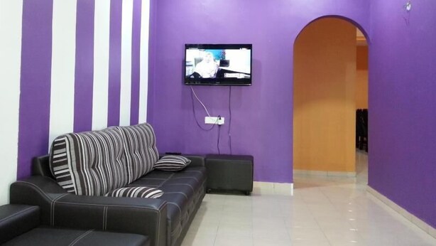 A living room with a purple wall, a black sofa with striped pillows, and a flat-screen TV mounted on the wall. - Noor Homestay (Kangar)