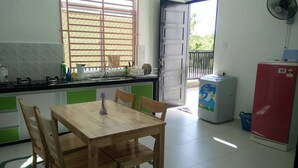 A compact kitchen with a dining table and chairs, a refrigerator, a washing machine, and a window with blinds. - Noor Homestay (Kangar)