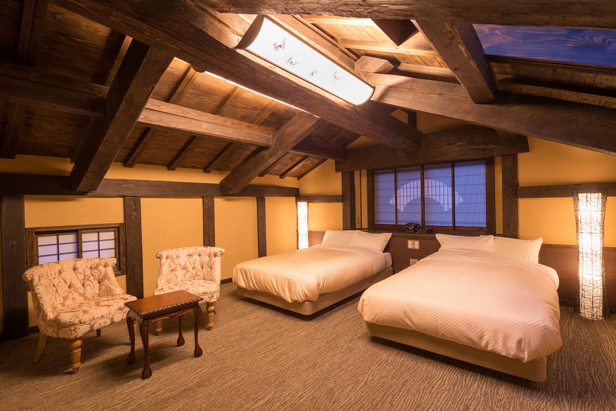 Una habitación con techo inclinado, vigas de madera, dos camas, una silla y una mesa.