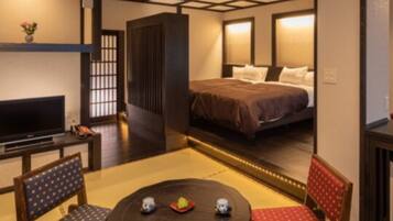 A traditional Japanese-style room with tatami flooring, a low table with cushions, a bed with a dark headboard, and a television on a stand.