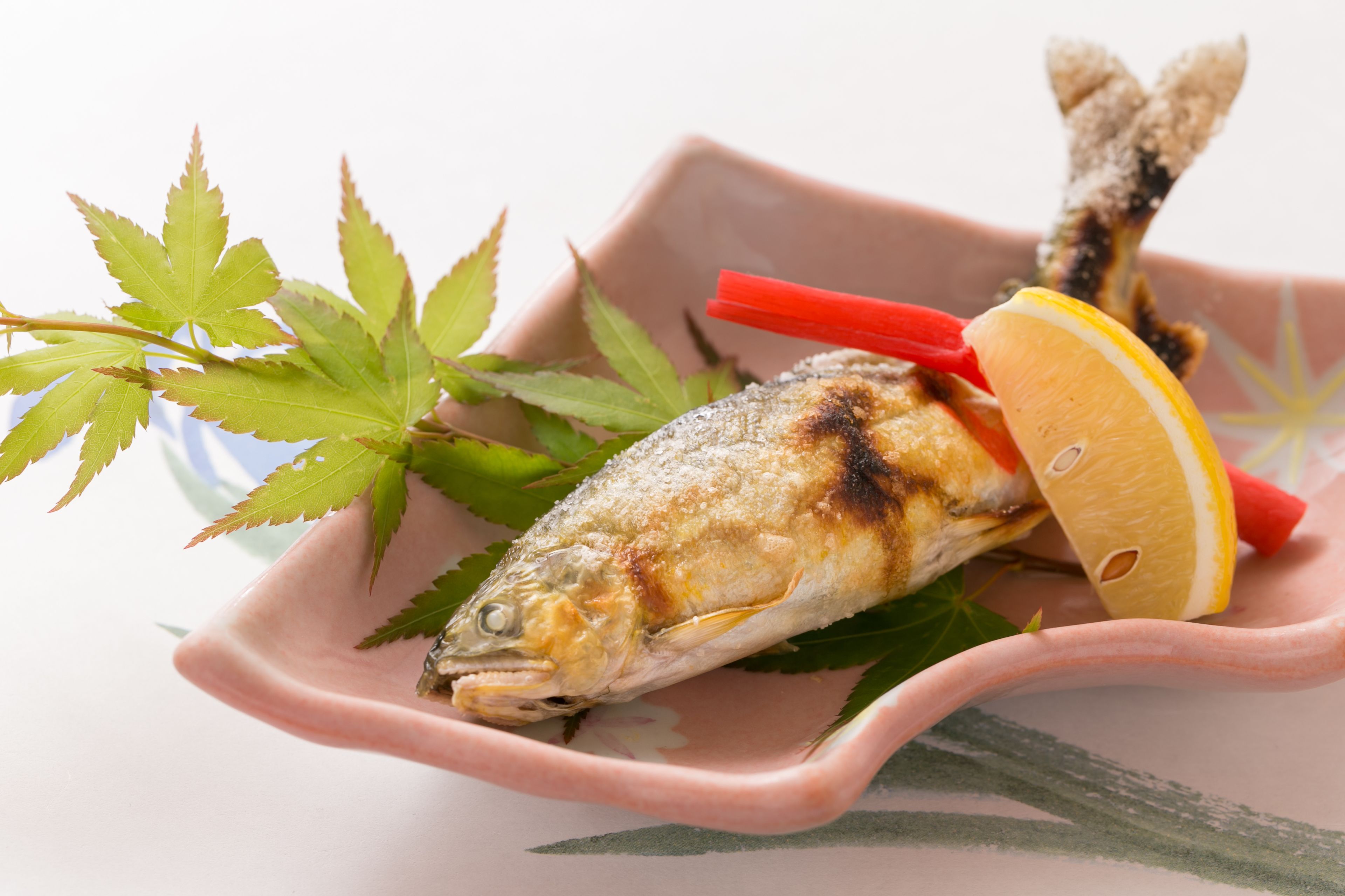 Ein gegrillter Fisch auf einem Teller mit Zitronenscheiben und grünen Blättern.