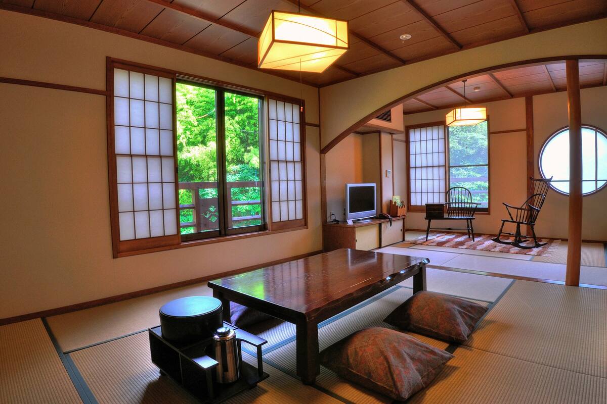 Una habitación japonesa tradicional con suelo de tatami, una mesa baja, cojines, un televisor y puertas correderas con vistas a zonas verdes.