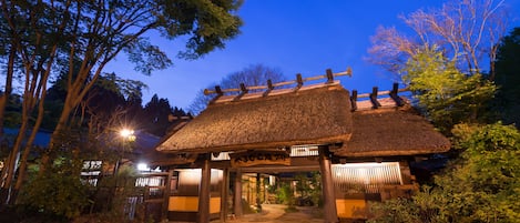 A traditional Japanese building with a thatched roof and wooden pillars, surrounded by trees and a paved entrance.