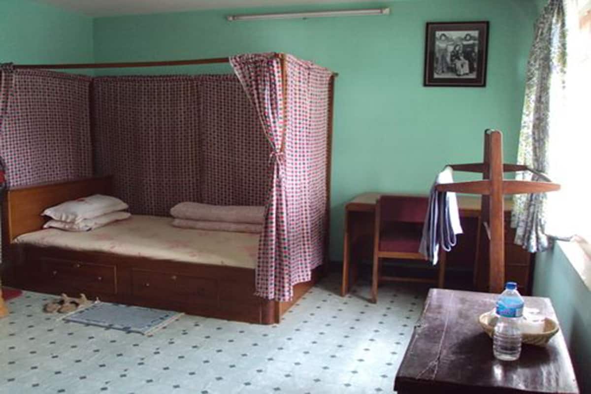 A single bed with a checkered canopy, a small table with a water bottle, and a chair.