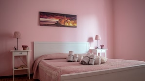 Un lit bien fait, avec des serviettes pliées, une tête de lit blanche et un mur rose orné d’un tableau représentant un coucher de soleil.