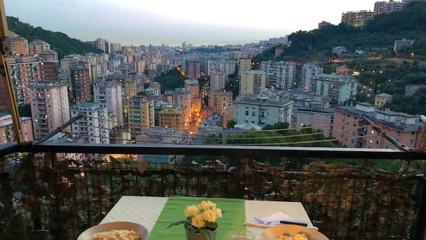 Un balcon avec une table dressée pour un repas, offrant une vue sur un paysage urbain dense, avec des bâtiments et une colline en arrière-plan.