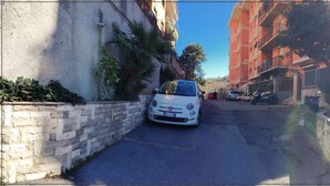 Une rue étroite où est garée une voiture blanche sur le bord, entourée de bâtiments et de verdure.