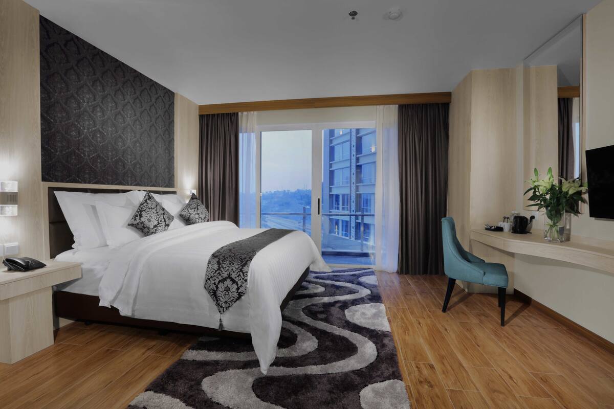 A modern hotel room with a large bed, a desk with a chair, a television, and a view of a building through the window.