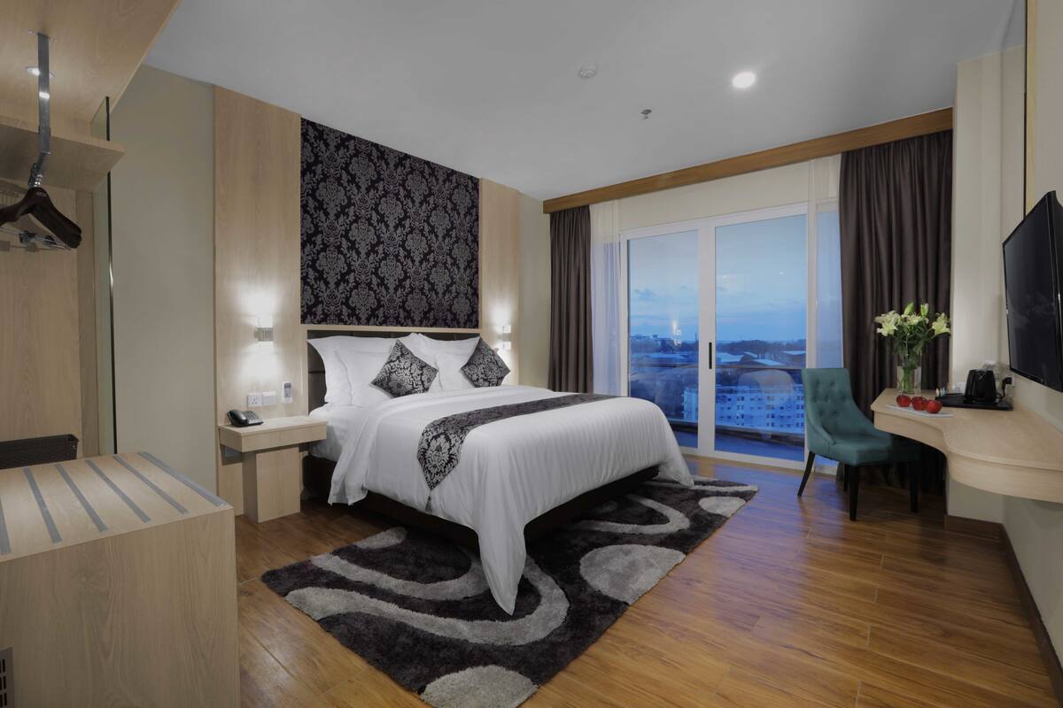 A modern hotel room with a large bed, a desk with a chair, a TV, and a view of the city.