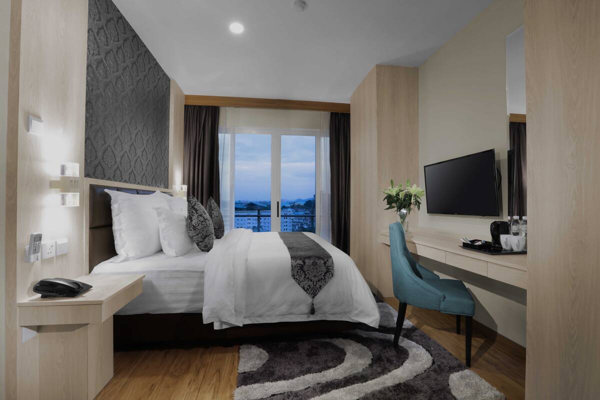 A modern hotel room with a bed, a desk, a chair, a TV, and a view of a cityscape.