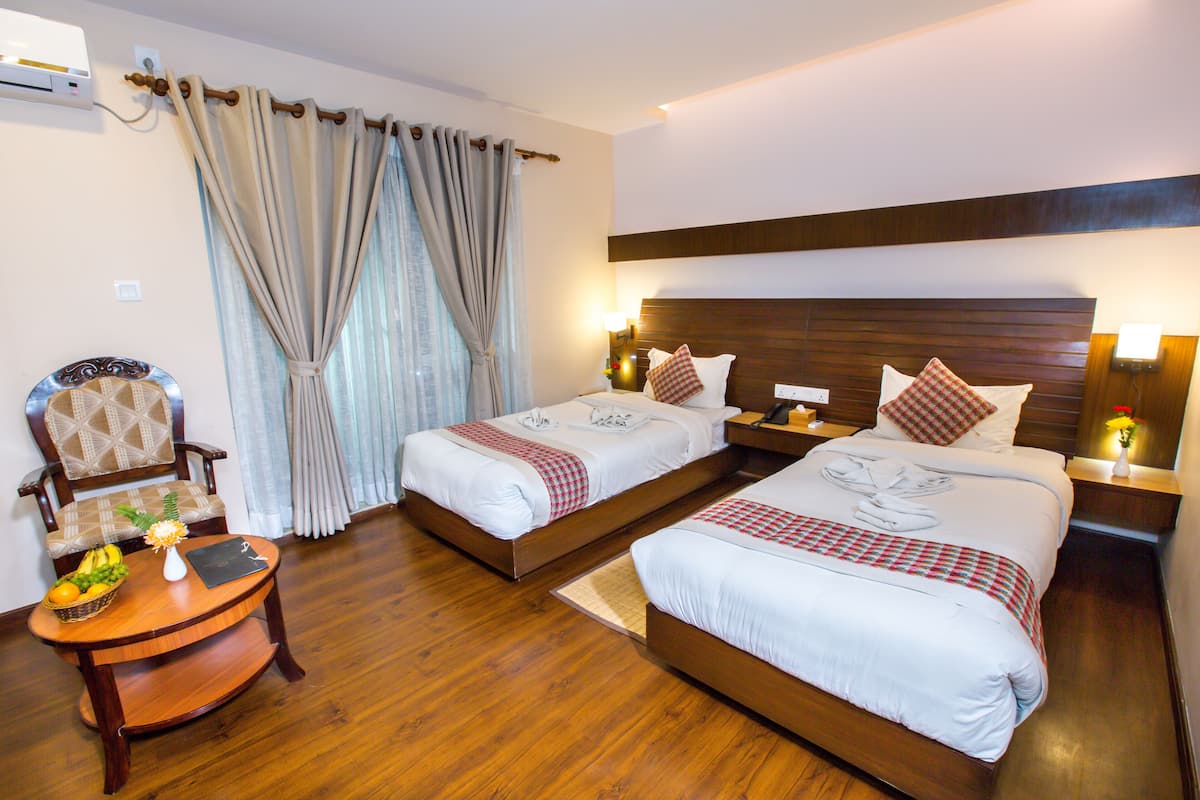 A hotel room with two beds, a wooden chair, a small table with fruits and a vase, and a window with curtains.